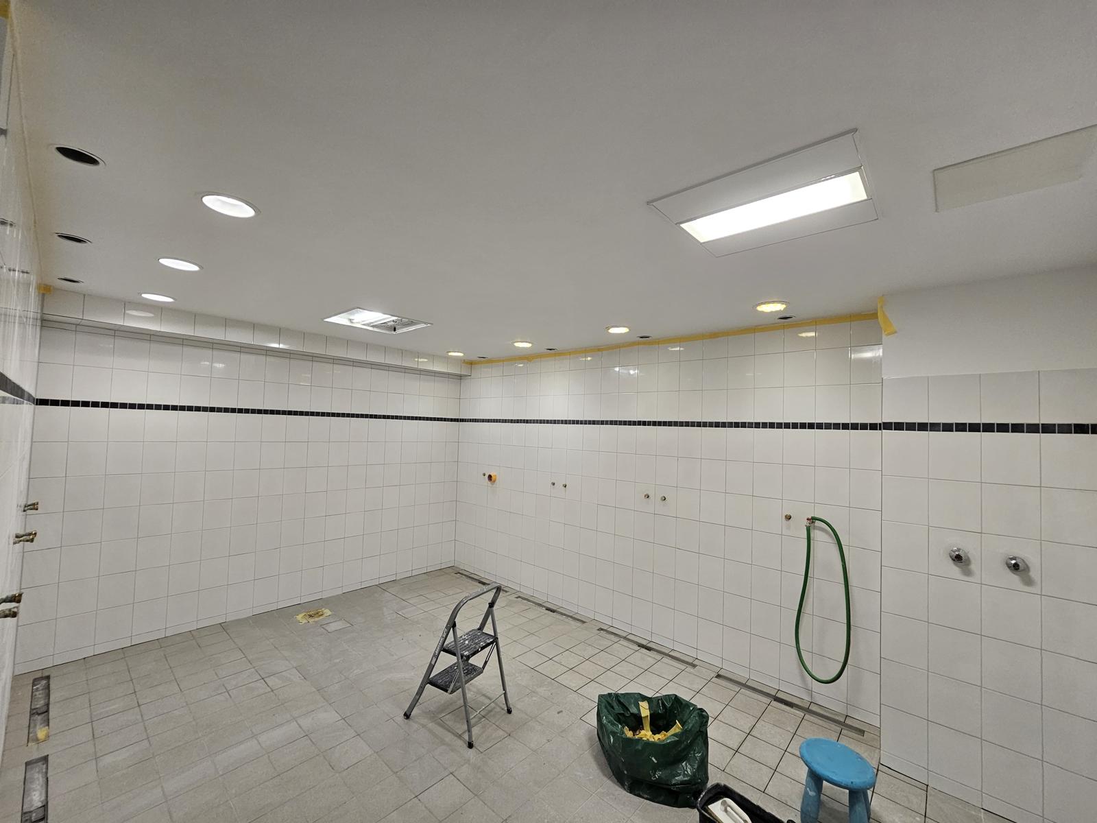 Der große Sanitärraum fertig gefliest mit schwarzer Zierbordüre und neu gestrichener Decke; die Anschlüsse für die Waschtischreihe stehen bereit.