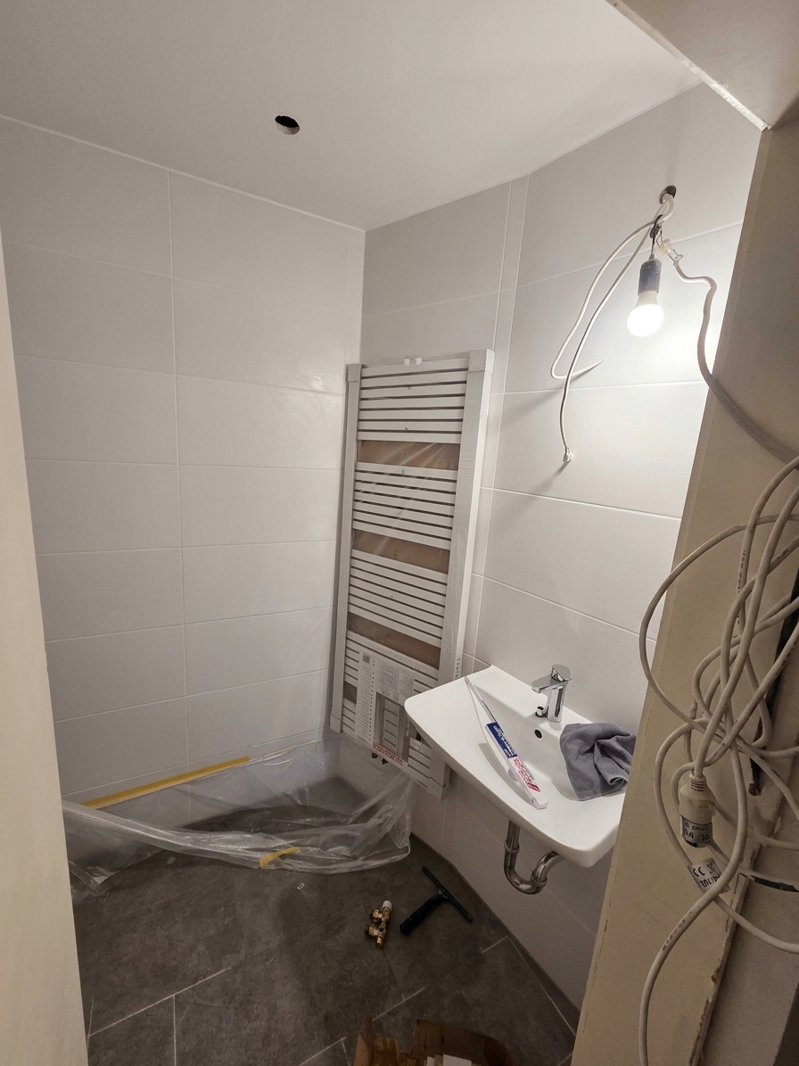 Fast fertig: neue Wandfliesen, dunkler Bodenbelag, montierter Handtuchheizkörper und neues Waschbecken – der Duschbereich ist noch abgeklebt.
